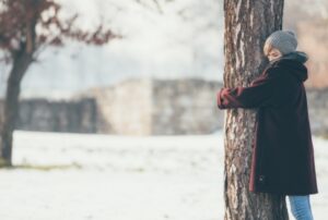 a woman in a burgundy coat hugs a tree in snowy field